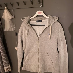 Grå hoodie från Polo Ralph Lauren - Säljer en klassisk grå hoodie från Polo Ralph Lauren. Den har en dragkedja framtill och en broderad logga på bröstet. Perfekt för en avslappnad stil. Den är lite för stor för M men de kan gå bra.