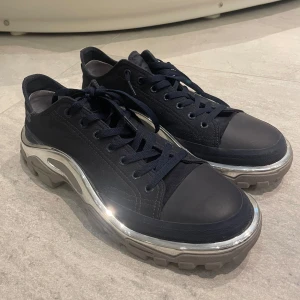 Svarta sneakers från Adidas x Raf Simons - Snygga svarta sneakers från Adidas i samarbete med Raf Simons. Skorna har en unik design med en robust sula i silver och grått, samt snörning framtill. Perfekta för dig som vill ha en stilren men ändå iögonfallande look.