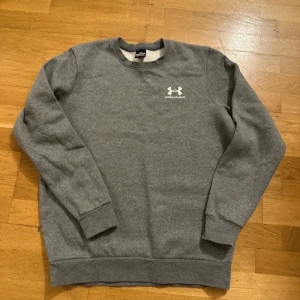 Under armour  - Säljer en stilren grå sweatshirt från Under Armour med deras logga tryckt på bröstet. Tröjan har en klassisk rund hals och långa ärmar, perfekt för en avslappnad look. Passar bra till både träning och vardag.