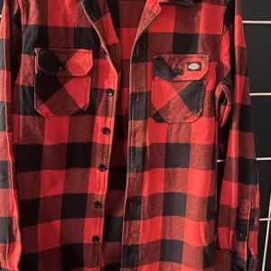 Röd och svart rutig skjorta från Dickies - Snygg röd och svart rutig skjorta från Dickies med klassisk design. Skjortan har långa ärmar och knappar framtill samt två bröstfickor med lock. Perfekt för en avslappnad stil.