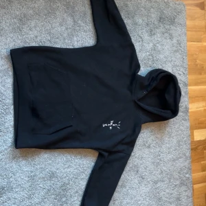Svart Travis Scott hoodie - Säljer en svart Travis Scott hoodie med cactus jack broderat på bröstet och ett stort tryck på ryggen med texten 'LOOK MOM I CAN FLY'. Hoodien har en klassisk känguruficka och en justerbar huva. Perfekt för dig som gillar streetwear och unika tryck! Hör av er vid fler frågor 
