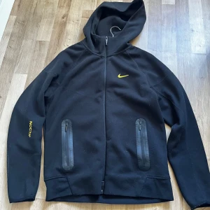 Svart jacka från Nike NOCTA - Säljer en stilren svart jacka från Nike NOCTA med dragkedja och huva. Jackan har gula detaljer och två praktiska fickor med dragkedjor framtill. Perfekt för en sportig look.