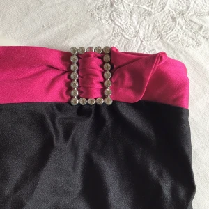 Svart och rosa tubtopp med strassdetalj - Snygg svart tubtopp med en bred rosa kant upptill och en glittrande strassdetalj. Perfekt för en kväll ute eller en festlig tillställning. Toppen är axelbandslös och har en elegant design.