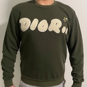 Olivgrön tröja  - Säljer en olivgrön tröja med ett stort, vitt logotyptryck på framsidan och en liten broderad bi. Tröjan har långa ärmar och en rund halsringning. 