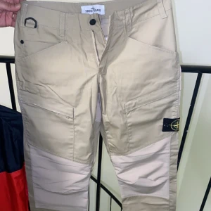 Beige cargopants från Stone Island - Snygga beige cargopants från Stone Island med flera praktiska fickor och en stilren design. Byxorna har en normal passform och är perfekta för en avslappnad stil. Märkesloggan syns tydligt på sidan. Och den är äkta 