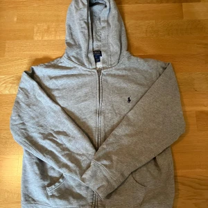 Grå hoodie från Polo Ralph Lauren - Säljer en klassisk grå hoodie från Polo Ralph Lauren med dragkedja och huva. Den har en broderad logga på bröstet och är tillverkad i mjukt material för extra komfort. Perfekt för en avslappnad stil.