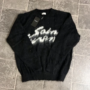 Svart tröja från Saint Laurent - Säljer en svart tröja från Saint Laurent med vit text på framsidan. Tröjan har långa ärmar och en rund halsringning. Perfekt för en stilren look.
