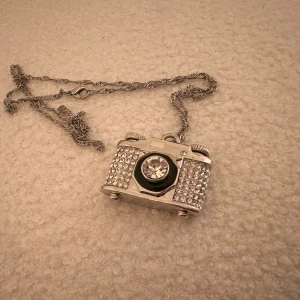 Kamerahalsband med strass - Unikt halsband med en kamerahänge dekorerat med glittrande strass. Hänget är i form av en klassisk kamera med en stor kristall i mitten. Kedjan är i silverfärg och har en enkel låsning. Perfekt för fotointresserade eller som ett statement-smycke.