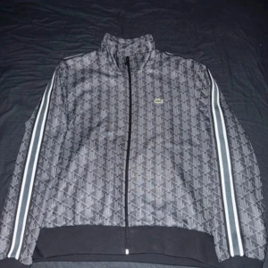 Lacoste monogram - Säljer en snygg  tröja från Lacoste i grått med vita ränder längs ärmarna. Tröjan är helt ny med prislapp och är i storlek XL men passar även L. Tröjan har dragkedja och en diskret logga på bröstet. Perfekt för en sportig look! 