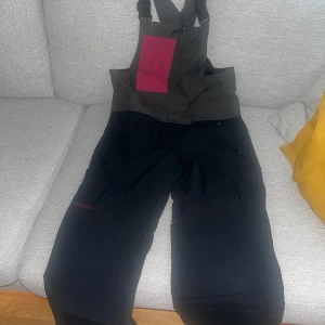 Svarta täckbyxor med hängslen från Airblaster - Säljer ett par svarta täckbyxor med hängslen och dragkedjor. Byxorna har en praktisk ficka på bröstet i röd färg och är perfekta för vinterns kalla dagar. De är designade för att hålla dig varm och torr med högkvalitativt material. Jag brukar använda dom för att åka snowboard. Inga snö kom in och jag älskar ventilation dragkedjor