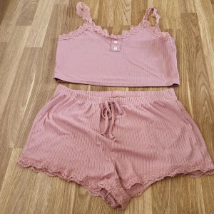 Pyjamasset - Säljer en söt rosa ribbad linnetopp med spetsdetaljer och knappar framtill. Med tillhörande shorts. Toppen är ärmlös och har en croppad passform. Perfekt för varma dagar eller mysiga hemmakvällar. Tillverkad i mjuk polyester.