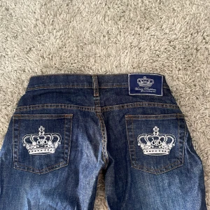 Blå Victoria Beckham jeans  - Snygga blå jätte fräcka omsydda Victoria Beckham jeans med broderade kronor på bakfickorna. Jeansen jätte low wested. har en klassisk femficksdesign och en knappgylf. Perfekta för en stilren look.