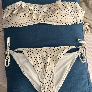 Prickig bikini med volangdetaljer - Vit bikini med svarta prickar och volangdetaljer på toppen. Bikinin har knytband på sidorna av trosorna för justerbar passform. 