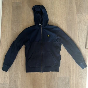 Mörkblå huvtröja från Lyle & Scott - Säljer en snygg mörkblå huvtröja från Lyle & Scott med dragkedja och en gul broderad logga på bröstet. Tröjan har långa ärmar och en praktisk huva med snörning. Perfekt för en avslappnad stil.