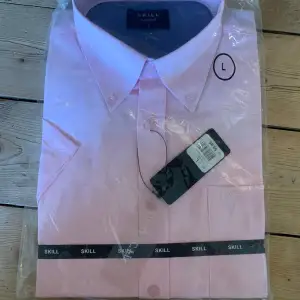 Säljer en stilren ljusrosa skjorta från Skill. Skjortan har en klassisk krage och är kortärmad med knappar framtill. Perfekt för en elegant look. Aldrig använd.
