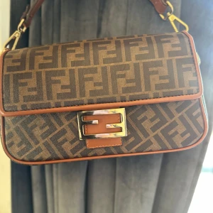 Fendi väska  - Snygg brun axelväska med monogrammönster och gulddetaljer. Väskan har en justerbar axelrem och ett elegant spänne framtill. Perfekt för att ge din outfit en lyxig touch.