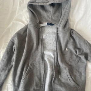 Grå hoodie från Ralph Lauren - Jag säljer en super fin grå zip up hoodie från Ralph lauren! Har ett klassiskt vit broderad Ralph lauren logotyp där fram som visar att den är äkta. Har använts några gånger men är i bra skick. Pris kan diskuteras.