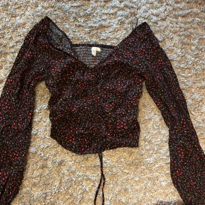 Blommig svart blus med långa ärmar - Säljer en söt svart blus med blommigt mönster i rött, lila och gult. Blusen har långa ärmar och en smickrande v-ringning med knytning framtill. Den är välanvänd men ser fortfarande ny ut