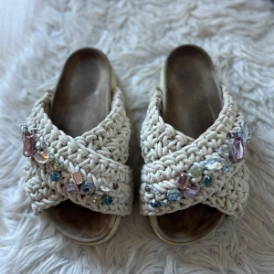 Inuikii tofflor med stenar - Snygga beige iniukki tofflor med korsade remmar i stickat material och glittrande pärldetaljer. Perfekta för en somrig look. Sandalerna har en öppen tå och en bekväm sula. 