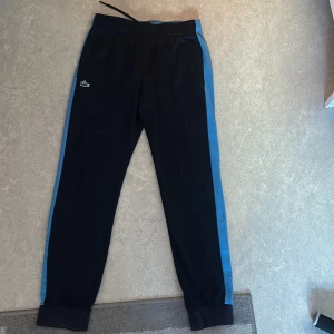 Mörkblå/blå träningsbyxor från Lacoste Sport - Snygga svarta träningsbyxor från Lacoste Sport med blå och vita detaljer längs sidorna. Perfekt för en chill dag ute