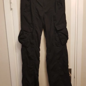 Nya Cargobyxor svarta strl 38 från H&M - Snygga svarta cargobyxor med flera praktiska fickor och knappstängning. Perfekta för en avslappnad stil med en touch av funktionalitet. Byxorna har en elastisk midja för extra komfort.