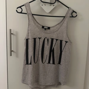 Grå linnetopp med texten 'LUCKY' - Snygg grå linnetopp från Divided med stor svart text 'LUCKY' på framsidan. Perfekt för en avslappnad stil. Tillverkad i mjukt material för extra komfort.