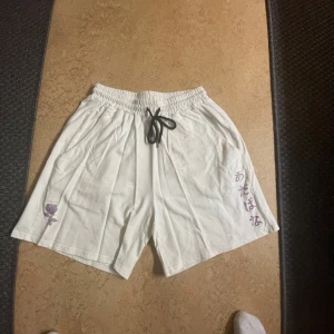 Vita shorts med japansk text - Säljer ett par vita shorts med elastisk midja och svart snörning. De har en unik detalj med japansk text och en liten lila ros på ena benet. Perfekta för en avslappnad stil.