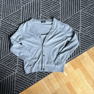 Grå kofta från Zara Man - Säljer en stilren grå kofta från Zara Man. Koftan har en v-ringad design med knappar framtill och långa ärmar. Perfekt för lager-på-lager under kallare dagar. Passar både till vardags och mer uppklädda tillfällen.