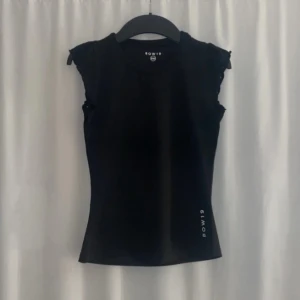 Svart topp från Intersport - Snygg svart träningströja från Intersport med korta ärmar och en tight passform. Toppen har volangdetaljer vid ärmarna. Perfekt för en trendig look! Nypris är 420kr