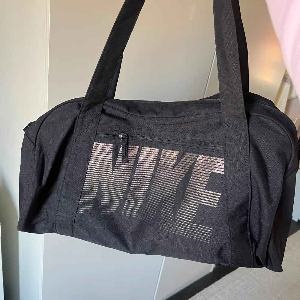 Säljer en svart axelväska från Nike med stort logotyptryck på framsidan. Väskan har en dragkedja och långa axelremmar, perfekt för att bära med sig allt du behöver. Praktisk och stilren design för alla tillfällen.. Laukut & Käsilaukut.