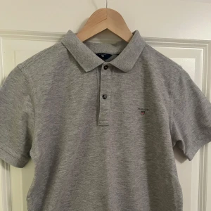 Grå pikétröja från Gant - Säljer en stilren grå pikétröja från Gant med klassisk krage och knappar framtill. Tröjan har en liten broderad logga på bröstet och är kortärmad. Perfekt för en avslappnad men snygg look.