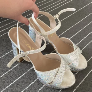 Vita glittriga sandaler med klack - Snygga vita sandaler med öppen tå och klack. Skorna är dekorerade med glittriga stenar och har en justerbar rem runt ankeln. Perfekta för att ge en extra touch av glamour till din outfit.