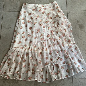 Vit blommig kjol med volang - Säljer en superfin vit kjol från Vila med blommigt mönster i rosa och grönt. Kjolen har en omlott-design med volanger som ger en romantisk känsla. Perfekt för sommarens alla tillfällen!
