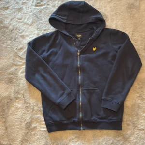 Mörkblå hoodie från Lyle & Scott - Säljer en stilren mörkblå hoodie från Lyle & Scott med dragkedja och huva. Tröjan har ett litet gult emblem på bröstet och är perfekt för en avslappnad stil. Passar bra för kyligare dagar.