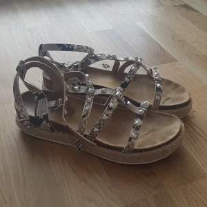 Ormskinnsmönstrade sandaler från Steve Madden - Snygga sandaler från Steve Madden med ormskinnsmönster och nitar. De har flera remmar och en bekväm sula i naturmaterial. Perfekta för en trendig look i sommar.