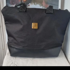 Svart axelväska med gulddetaljer - Snygg svart axelväska med gulddetaljer och dragkedja. Väskan har en stilren design med en praktisk ficka på baksidan. Perfekt för att bära med sig det nödvändigaste med stil. Se bilder för vart den är sönder,går att laga