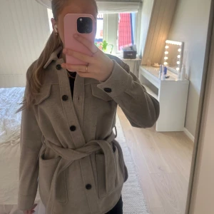 Beige overshirt från Dobber - Säljer en stilren beige overshirt från Dobber i storlek XS. Den har långa ärmar och knäpps med knappar framtill. Den har en knytdetalj i midjan och två bröstfickor med lock. Använd ett fåtal gånger.