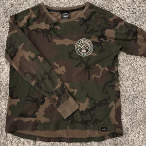 Camouflage långärmad tröja från Dope - Säljer en cool långärmad tröja från Dope med ett klassiskt camouflage-mönster. Tröjan har en rund halsringning och ett stort tryck på ryggen med Dope-loggan. Perfekt för en avslappnad stil.