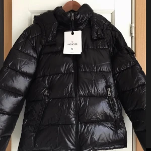 Svart dunjacka från Moncler - Säljer en snygg svart dunjacka från Moncler. Jackan har en glansig finish och är utrustad med dragkedja och huva. Perfekt för kyliga dagar med stil. Moncler-logga på ärmen ger en exklusiv touch.