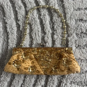 Guldig kuvertväska med pärlhandtag - Elegant guldig kuvertväska dekorerad med paljetter och blommönster. Väskan har ett pärlhandtag som ger en lyxig känsla. Insidan är klädd i satin för en mjuk och fin finish.
