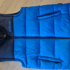 Blå dunväst från Napapijri - Snygg blå dunväst från Napapijri med dragkedja framtill. Västen har en stilren design med två praktiska fickor. Perfekt för kyligare dagar när du vill hålla värmen utan att kompromissa med stilen.