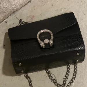 Svart axelväska med kedja - Snygg svart axelväska i krokodilmönstrat material med en elegant kedja som axelrem. Väskan har ett dekorativt spänne framtill och en röd insida med dragkedja för extra säkerhet. Perfekt för att ge din outfit en stilfull touch.