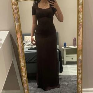 Brun långklänning med snörning - Elegant brun långklänning med korta ärmar och en snygg snörning i midjan. Klänningen har en ribbad textur som ger en stilren look. Perfekt för en kväll ute eller en speciell tillställning.