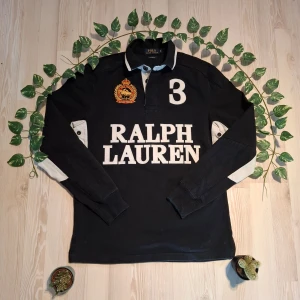 Svart rugbytröja från Ralph Lauren - Säljer en svart rugbytröja från Ralph Lauren med stora vita bokstäver och siffran 3 på både fram- och baksidan. Tröjan har långa ärmar och en broderad detalj på bröstet. Perfekt för en stilren look!