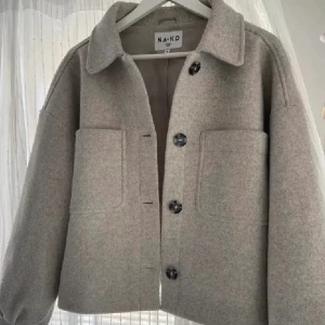Beige jacka från NA-KD - Säljer en stilren beige jacka från NA-KD med stora knappar och två framfickor. Jackan har en klassisk krage och en avslappnad passform, perfekt för höst och vår. Den är långärmad och tillverkad i ett mjukt material.
