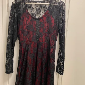 Vinröd spetsklänning från Gothicana by EMP - Snygg vinröd klänning med svart spets från Gothicana by EMP. Klänningen har långa spetsärmar och ett vackert blommönster. Den är dekorerad med knappar framtill och har en elegant design som passar perfekt för speciella tillfällen.