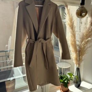 Beige kappa med bälte - Elegant beige kappa med bälte i midjan. Perfekt för en stilren look. Långärmad design med klassisk krage🥰