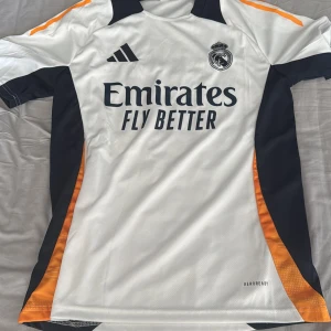 Real Madrid tränings tröja från Adidas - Snygg real madrid tränings tröja från Adidas med svarta och orange detaljer. Tröjan har korta ärmar och klubbmärke på bröstet. Perfekt för fotbollsfans som vill visa sitt stöd. Tillverkad i ett lätt och andningsbart material. Stl är M