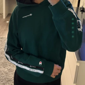 Grön hoodie från Champion - Säljer en snygg grön hoodie från Champion med logotypdetaljer längs ärmarna och på bröstet. Tröjan har en klassisk känguruficka och ribbade muddar. Perfekt för en avslappnad stil.