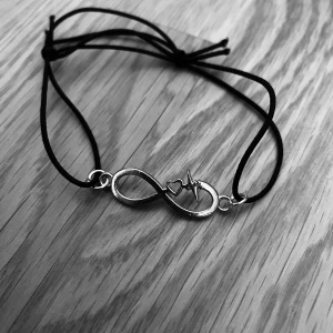 By prisförslag- Armband med guld oändlighetssymbol och hjärtslag - Snyggt armband med en oändlighetssymbol i guld och ett hjärtslag i mitten. Armbandet har en svart justerbar rem som gör det enkelt att anpassa storleken. Perfekt för att ge en personlig touch till din stil.